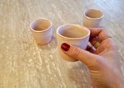 Vous&G agence immobilière Une main recouverte de vernis à ongles rouge tient une petite tasse en céramique sur une surface en marbre beige. Deux tasses similaires sont posées sur la table à l'arrière-plan.
