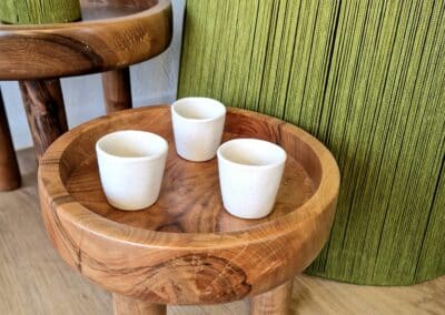 Vous&G agence immobilière Une petite table ronde en bois avec trois pieds courts contient trois tasses en céramique blanche. La table est posée sur un sol en bois clair, avec en arrière-plan de grands objets verts et texturés en bois.