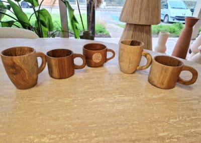 Vous&G agence immobilière Cinq tasses en bois munies d'une anse sont disposées en rang sur une table de couleur claire, avec en arrière-plan de la verdure, des poteries et une fenêtre montrant une voiture en stationnement.