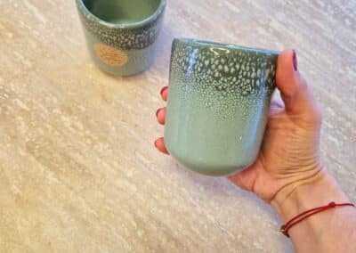 Vous&G agence immobilière Une main recouverte de vernis à ongles rouge tient une tasse en céramique verte mouchetée ; une autre tasse similaire est posée sur une surface texturée de couleur claire à l'arrière-plan.