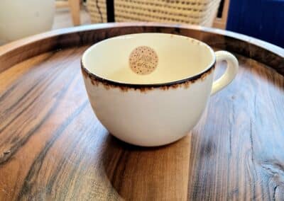 Vous&G agence immobilière Une tasse en céramique blanche avec un bord marron est posée sur un plateau rond en bois. La tasse contient un petit disque en bois sur lequel figure un texte. L'arrière-plan comprend un panier tressé avec des cœurs rouges et des meubles en bois.