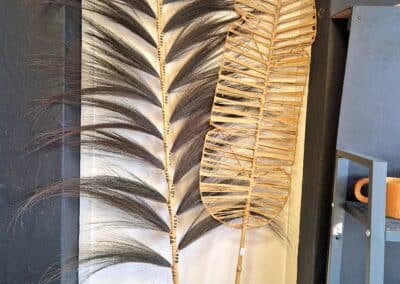 Vous&G agence immobilière Deux grandes feuilles de palmier décoratives, l'une avec de fines folioles foncées et l'autre en tissu clair, sont appuyées contre un mur blanc à côté d'une étagère noire.