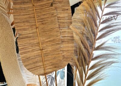 Vous&G agence immobilière De hautes feuilles de palmier séchées et des herbes sont disposées dans un présentoir décoratif, avec une chaise tissée et un papier peint abstrait en arrière-plan, créant ainsi une scène de décoration intérieure naturelle et bohème.